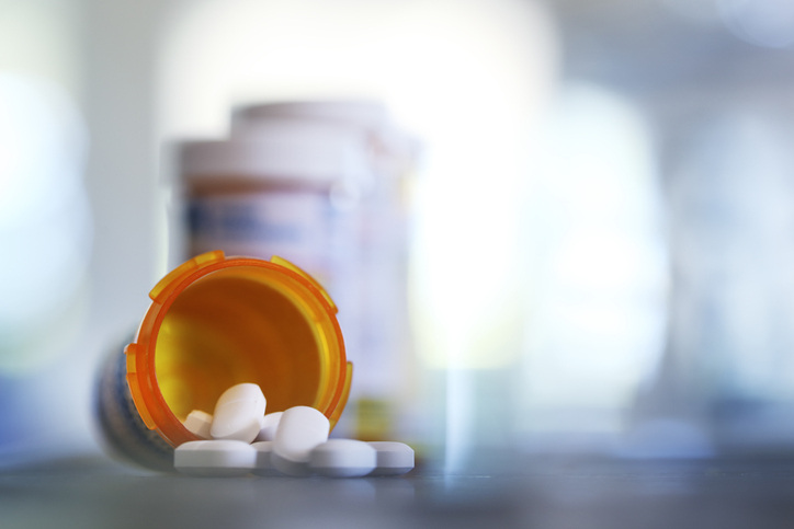 Pills pour out of a prescription medication bottle onto a kitchen counter. Several other pill bottles stand out of focus in the background.