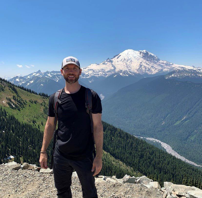 Man in dark clothing and baseball cap stands in front of mountain landscape