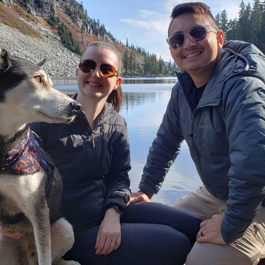 couple poses with their dog in front of lake and mountain landscape