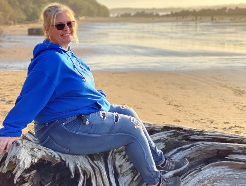 blonde woman sitting on fallen tree and taking photo with coast in the background
