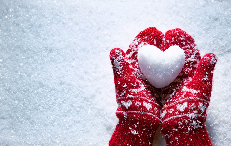 Female hands in knitted mittens with snowy heart against snow background. Love, winter and Valentines day romantic creative concept with copy space for text