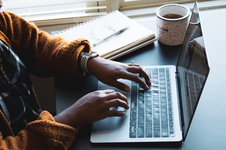 Black woman typing on her laptop with documents and coffee beside her