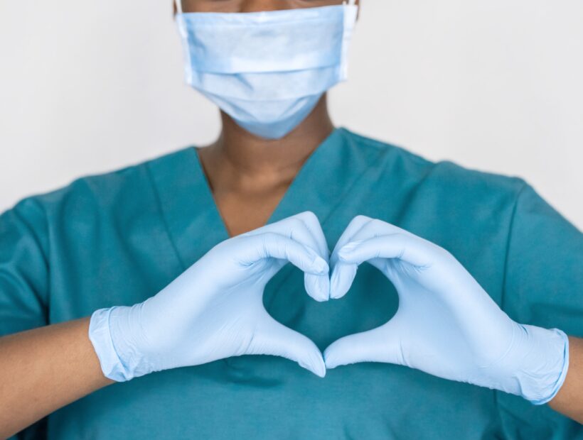 Female african professional medic nurse wear face mask, gloves, blue green uniform showing heart hands shape. Medical love, care and safety symbol, corona virus health protection sign concept. Closeup