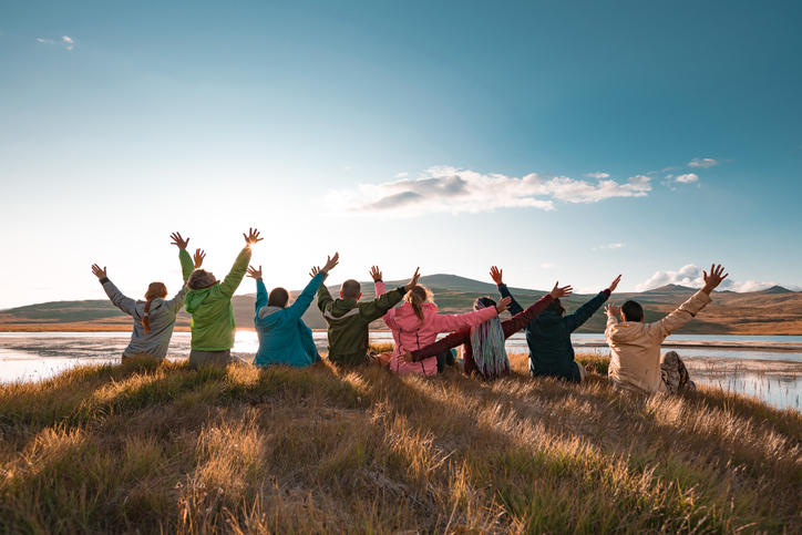 Big group of happy tourists are sitting with raised arms at beautiful sunset lake and mountains