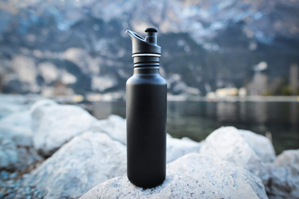 Close-up black of steel eco thermo water bottle on the background of the lake in the mountains. Copy space. Zero waste, no plastic, sustainability.